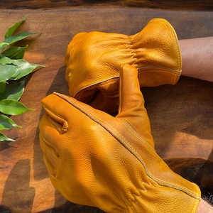 May include: A pair of yellow leather gloves with contrasting stitching detail. The gloves are folded in half and the stitching is visible on the outside of the gloves.