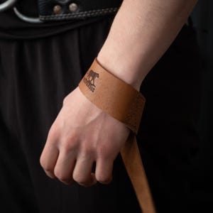 May include: Brown leather wrist strap with a stitched design and a black panther logo with the text "UNAPOLOGETIC" engraved on it.