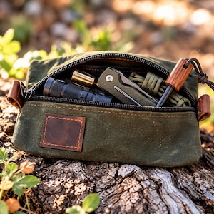 May include: An olive green waxed canvas zipped pouch featuring a brown leather patch on the front. The open pouch contains a black torch, a metallic brass-coloured object, a folding pocket knife, and a coil of olive green paracord.