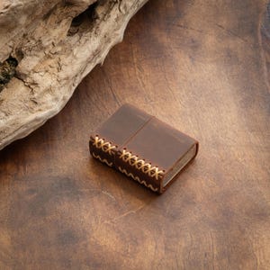 May include: A brown leather lighter case with a stitched design. The case is closed and has a worn look.
