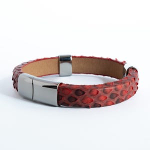 May include: Red snakeskin bracelet with a magnetic clasp. The bracelet features a textured, scaly pattern in shades of red and brown. The clasp is made of polished silver-tone metal. The interior of the bracelet is a light brown color.