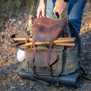 May include: A brown leather and green canvas rucksack with a large flap and multiple pockets. The rucksack is filled with various items, including a wooden stick, a white object, and a brown leather strap.