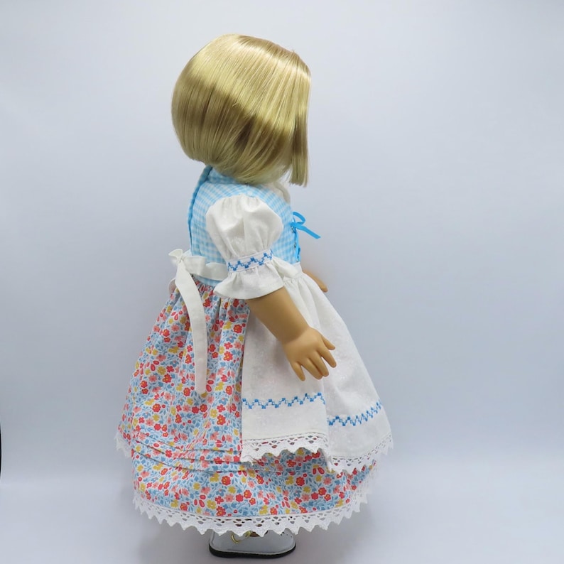 May include: A doll wearing a blue and white gingham dress with white sleeves and a floral print skirt. The dress has a white apron with blue embroidered details and a white lace trim. The doll has blonde hair.