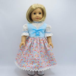 May include: A doll wearing a floral print dress with a blue gingham bodice and white sleeves. The dress features a white lace trim and a blue ribbon detail. The doll has blonde hair and is wearing white shoes.