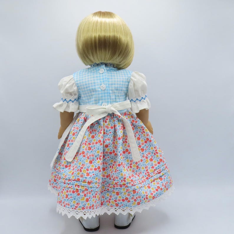 May include: A doll wearing a blue and white gingham dress with a floral print skirt. The dress has white sleeves, a white apron, and a white lace trim. The doll has blonde hair and is wearing white shoes.