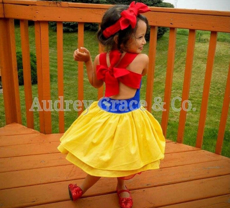 May include: A young girl wearing a Snow White costume. The dress is yellow with a blue bodice and a red bow on the back. She is wearing a red headband with a bow on top.