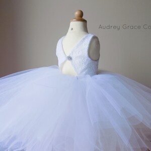 Op de afbeelding: Witte tulejurk met een kanten lijfje en een knoopsluiting met een juweel. De jurk heeft een volle rok en is perfect voor een speciale gelegenheid. Audrey Grace Co.