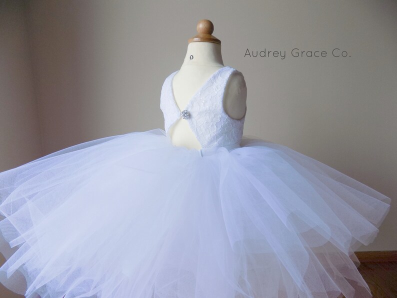 Op de afbeelding: Witte tulejurk met een kanten lijfje en een knoopsluiting met een juweel. De jurk heeft een volle rok en is perfect voor een speciale gelegenheid. Audrey Grace Co.