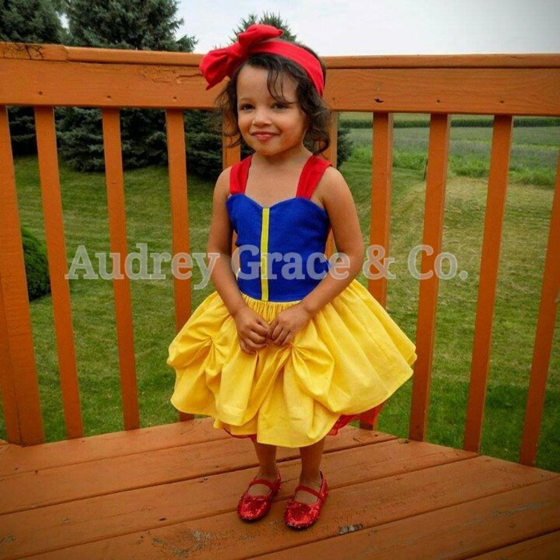 May include: A young girl dressed as Snow White, wearing a blue and yellow dress with a red bow headband. She is standing on a wooden deck.