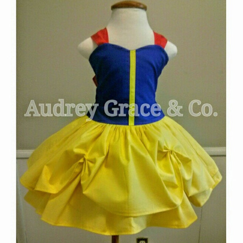 May include: A yellow and blue dress with a red bow. The dress has a yellow skirt with ruffles and a blue bodice with a yellow trim. The dress is on a white dress form.