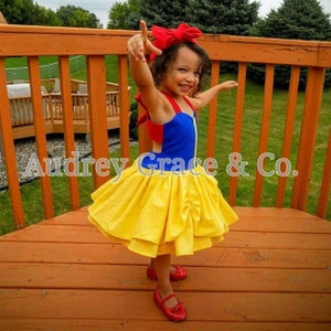 May include: A young girl wearing a Snow White costume. The dress is blue, red, and yellow with a white petticoat. She is wearing a red bow in her hair and red shoes.