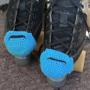 Puede incluir: Un par de patines de ruedas negros con cubrededos de ganchillo azul. Los cubrededos tienen una línea negra en la parte delantera.