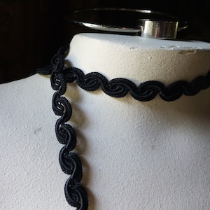 May include: A black, wavy trim with a scalloped edge is displayed on a white mannequin form. The trim is shown as a choker and a long strand.