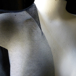 May include: A black mesh fabric garment draped over a white mannequin torso. The material features a fine, uniform hexagonal grid pattern that is semi-transparent, revealing the white surface of the mannequin underneath the dark textile.