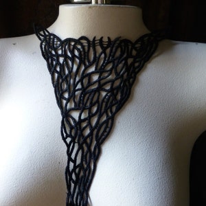 Op de afbeelding: Zwarte, open geweven ketting met een uniek, vloeiend ontwerp. De ketting wordt getoond op een mannequin, waarbij het ingewikkelde patroon en de vorm worden benadrukt. Het ontwerp lijkt te zijn gemaakt van een zacht, flexibel materiaal.