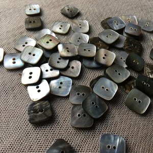May include: A collection of square, iridescent buttons with two holes each. The buttons vary in color from light gray to dark charcoal, scattered on a textured, neutral-toned surface. These buttons are ideal for sewing and craft projects.
