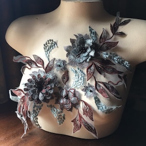 May include: A floral applique with intricate embroidery. The design features dark gray, burgundy, and light blue flowers and leaves. The applique is displayed on a mannequin, showcasing its potential use in fashion or craft projects.
