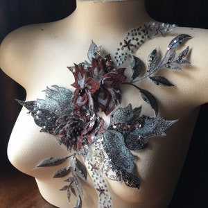 May include: A decorative floral applique with a cluster of burgundy and silver flowers and leaves. The applique is embellished with sequins and beads, and is displayed on a mannequin.
