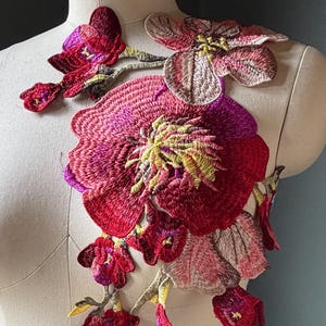 May include: A large floral embroidery applique featuring vibrant shades of red, magenta, and soft pink. The design consists of multiple layered flowers and buds with intricate stitching and yellow centers, arranged in a cascading vine-like pattern.