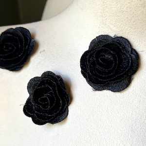 Puede incluir: Tres apliques de rosas de tela negra están dispuestos sobre una superficie blanca. Las rosas tienen diseños detallados de pétalos y están hechas de un material suave y texturizado. Los apliques son probablemente para manualidades o adornos.