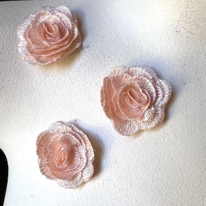Puede incluir: Tres apliques de rosas de tela rosa claro. Las rosas tienen un diseño detallado y en capas con un acabado texturizado. Se muestran sobre una superficie blanca, resaltando su color y textura suaves. Cada rosa mide unos 5 cm de diámetro.