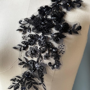 May include: A black floral lace applique featuring intricate embroidery, shimmering sequins, and small beads arranged in a botanical pattern on a sheer mesh backing. The design includes various flower shapes and leafy stems, creating a textured surface.