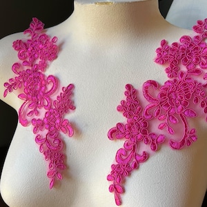 May include: Two fuchsia floral lace appliques are displayed on a white mannequin bust. The appliques feature intricate floral and swirling designs, ideal for embellishing garments or crafts. The lace is a vibrant, eye-catching colour.