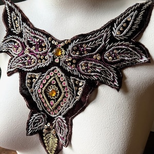 May include: An elaborate, decorative collar with a floral design. The collar is made of a dark burgundy velvet and is adorned with silver beads, sequins, and gold and pink accents. It is displayed on a white mannequin.
