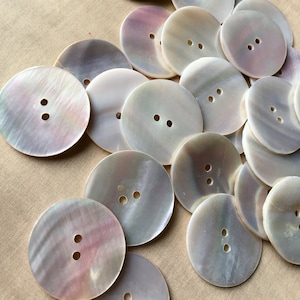 May include: A collection of iridescent, round buttons with two holes each. The buttons have a pearlescent sheen with hints of pink and purple, scattered on a tan surface. These buttons are ideal for sewing and craft projects.
