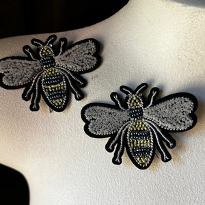 May include: Two embroidered bee brooches with black outlines, gray wings, and yellow and black striped bodies.