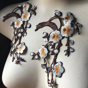 May include: Two white embroidered floral appliques with brown stems and yellow centres. The appliques are on a white background.