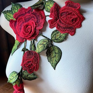 以下が含まれることがあります： 緑の葉が付いた刺繍された赤いバラのアップリケ。花のモチーフは3つのバラといくつかの葉を特徴とし、すべて詳細なステッチが施されています。アップリケは白いマネキンに表示され、刺繍の複雑な詳細を示しています。