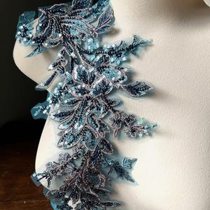May include: An elaborate teal and navy blue floral applique with sequin and bead embellishments. The applique is draped over a white mannequin, showcasing its intricate design and delicate details. The design features leaves and flowers.