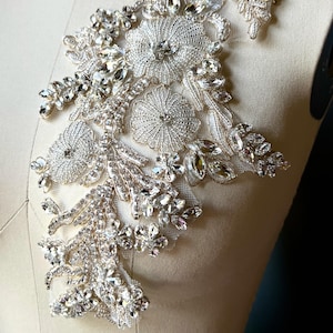 May include: A detailed, silver-toned applique featuring floral and leaf designs. The applique is heavily embellished with clear, sparkling rhinestones and beads, creating a luxurious and ornate appearance. The design is mounted on a cream-colored fabric.
