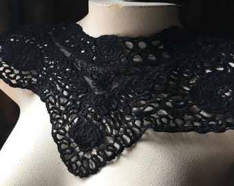 May include: A black lace collar with a floral design is displayed on a mannequin. The collar is made of delicate, open-weave lace and features several circular floral patterns. The collar is designed to be worn around the neck.