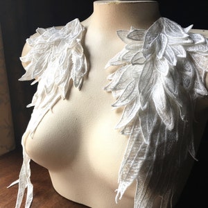 Puede incluir: Piezas de hombro de encaje blanco con diseño de plumas. El encaje delicado y detallado está dispuesto para parecer alas, con un efecto en capas. Las piezas se exhiben en un maniquí, mostrando la intrincada artesanía y textura de la tela.