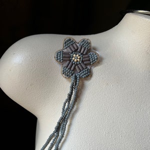 May include: A gray beaded flower with a long gray beaded tassel. The flower is made of small, round beads and has a center made of larger, brown beads.
