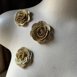 Puede incluir: Tres apliques de rosas doradas bordadas están dispuestos sobre un maniquí. Las rosas tienen patrones de pétalos detallados y están hechas de hilo metálico. El maniquí es de color beige neutro.