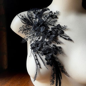 May include: Black floral lace applique with beaded accents. The applique features a cluster of black flowers with delicate leaves and stems. The applique is perfect for adding a touch of elegance to any garment.
