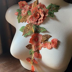 Puede incluir: Aplicación floral bordada en tonos coral, naranja, verde y marrón. La aplicación presenta flores y hojas detalladas, dispuestas en un diseño en cascada. El adorno floral se muestra en un maniquí.