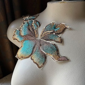 May include: A teal and gold embroidered applique in the shape of a butterfly. The applique is made of sequins and thread and has a delicate, feminine design.