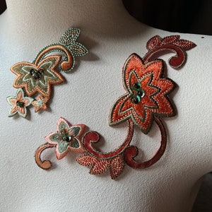 May include: Embroidered floral appliques in shades of orange, green, and gold. The appliques feature intricate detailing with beaded accents and are displayed on a mannequin. These decorative appliques are suitable for embellishing clothing or accessories.