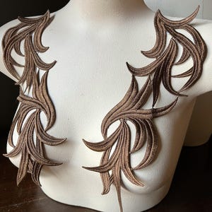 Op de afbeelding: Bruine geborduurde applicaties in een bladvormig ontwerp worden getoond op een witte mannequin. De applicaties zijn symmetrisch, waarbij elke kant de andere weerspiegelt. De gedetailleerde steken en kleur creëren een decoratief element.