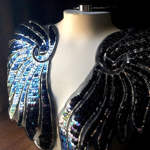 May include: Two iridescent blue and black sequined wings on a white mannequin. The wings are made of sequins and mesh fabric.