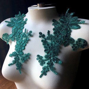 Könnte beinhalten: Zwei tealgrüne florale Spitzenapplikationen auf einer weißen Schaufensterpuppe. Die Applikationen bestehen aus zarter Spitze mit einem floralen Design.