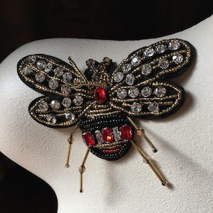 May include: A black and gold bee brooch with red rhinestone accents. The bee is embellished with beads and rhinestones.