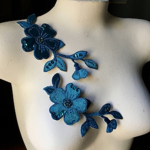 May include: Two teal floral appliques with sequin accents are displayed on a white mannequin. The appliques feature detailed flower and leaf designs, with a dark teal color and a touch of sparkle.