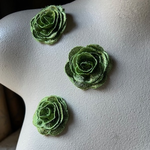 Puede incluir: Tres apliques de rosas de tela verde metálico. Las rosas están dispuestas sobre una superficie de tono neutro. Cada rosa tiene múltiples capas de pétalos, creando un efecto texturizado y dimensional. Los apliques son probablemente para manualidades o adornos.