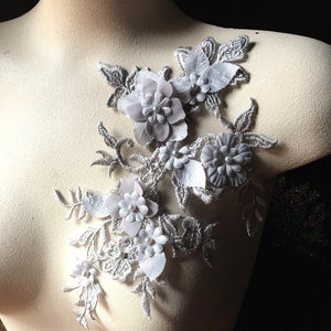 Op de afbeelding: Een zilveren kant-applicatie met witte bloemen van stof en parel accenten. De applicatie is perfect om een ​​toets van elegantie toe te voegen aan elk kledingstuk.