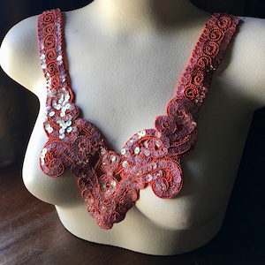 Puede incluir: Aplicación de color coral con cuentas y lentejuelas, exhibida en un maniquí. La aplicación presenta un diseño de cuello en V con intrincados patrones florales y lentejuelas brillantes. La pieza es probablemente para embellecer ropa o accesorios.